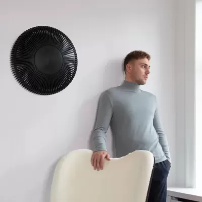 Homem com camisola cinzenta junto a cadeira creme e parede branca com decoração preta