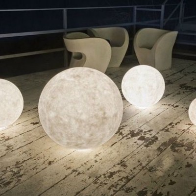 Quatro esferas luminosas brancas em vários tamanhos sobre chão de madeira com três cadeiras bege ao fundo