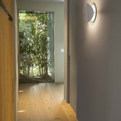 Luminária de parede branca semicircular com luz LED num corredor interior com chão de madeira e porta de vidro