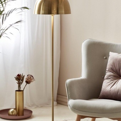 Candeeiro de pé dourado ao lado de cadeira cinzenta com almofada rosa, planta e cortina branca