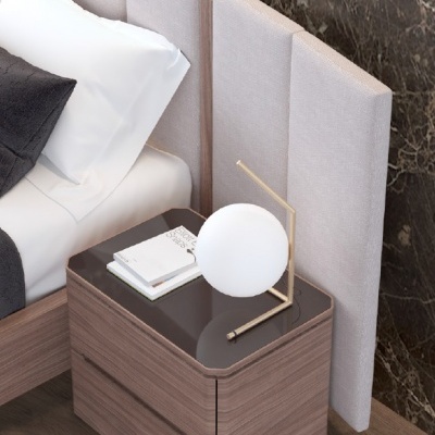 Mesa de cabeceira com abajur redondo branco e livros ao lado de cama com cabeceira bege