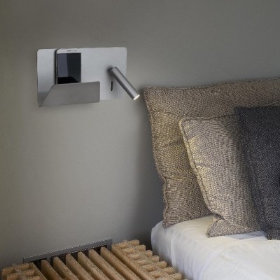 Suporte de parede metálico com lâmpada e telefone ao lado de cama com almofadas e aquecedor de madeira.