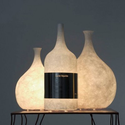 Três candeeiros em forma de garrafa translúcidos brancos com etiqueta preta sobre mesa metálica.