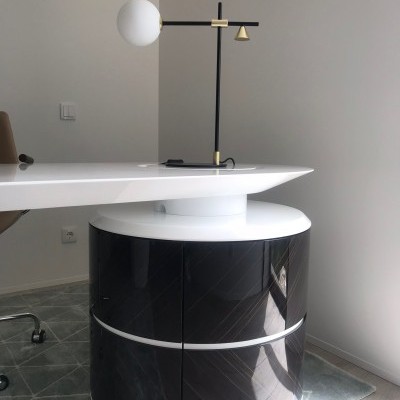 Mesa de escritório branca com apoio circular preto e branco, lâmpada moderna preta e dourada, cadeira castanha, tapete cinza e parede branca.