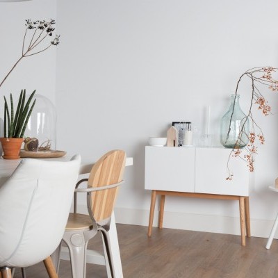 Sala de jantar moderna com cadeiras, mesa em madeira clara, plantas, móvel branco, garrafa com ramo e cabide com casaco