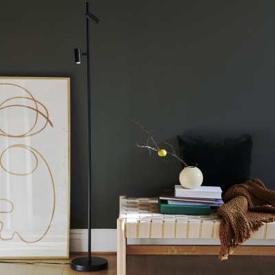 Banco branco com manta castanha, almofada e livros, lâmpada de pé preta e quadro com desenho abstrato encostado na parede cinza.