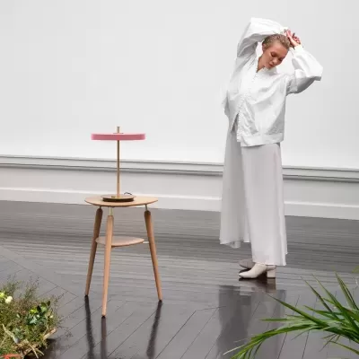 Mesa auxiliar em madeira clara com candeeiro de mesa de abajur rosa num piso escuro, numa sala minimalista
