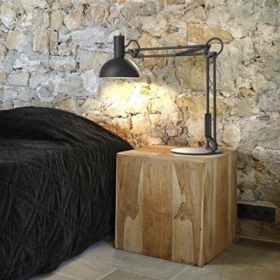 Candeeiro de mesa preto articulado sobre mesa de madeira natural junto a cama com edredão preto e parede de pedra