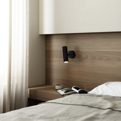 Abajur de parede preto numa cabeceira de madeira clara ao lado da cama com roupa cinza e branca