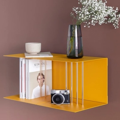 Estante amarela de metal com livros, câmara fotográfica, vaso com flores e recipiente cerâmico numa parede castanha