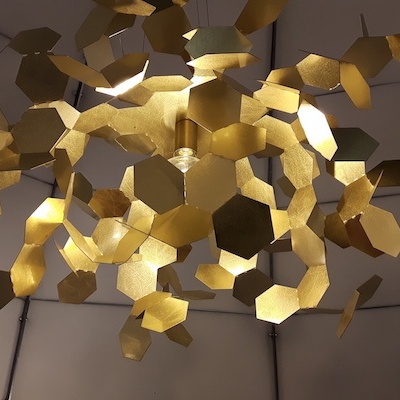 Candeeiro de teto dourado com peças hexagonais em teto falso branco.