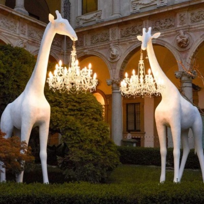 Esculturas brancas de girafas com candelabros pendurados no pescoço num jardim com edifício histórico ao fundo