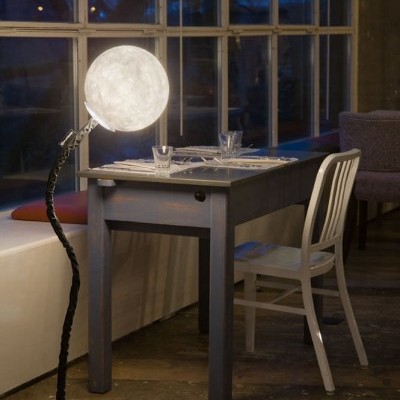 Lâmpada de pé com abajur redondo imitando a lua ao lado de mesa cinzenta e cadeira branca junto a uma janela grande à noite