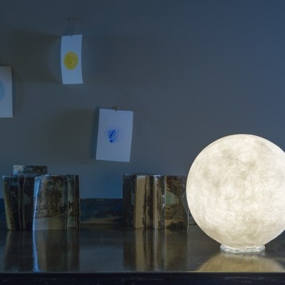 Abajur redondo branco iluminado sobre mesa metálica com objetos e desenhos ao fundo