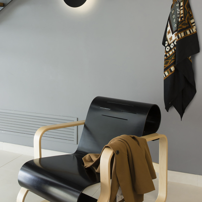 Cadeira moderna em madeira clara e couro preto com casaco castanho em ambiente minimalista cinza.