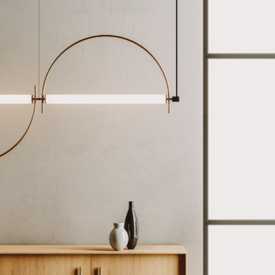 Candeeiro de parede dourado semicircular com luz linear, dois vasos decorativos sobre móvel de madeira, parede cinzenta e janela grande à direita.