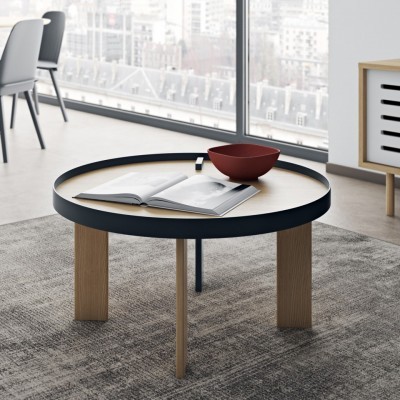 Mesa de centro redonda com tampo claro e borda preta, três pernas de madeira, taça vermelha e livro aberto sobre tapete cinza