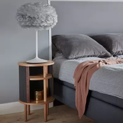 Mesa de cabeceira de madeira com porta preta ao lado de cama com roupa cinzenta e abajur branco
