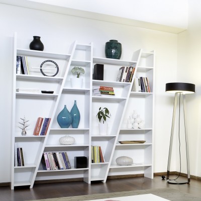 Estante branca inclinada com livros, vasos e candeeiro preto ao lado