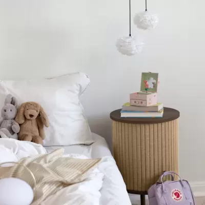 Cama branca com peluches e almofada ao lado de mesa redonda com livros e luminárias pendentes