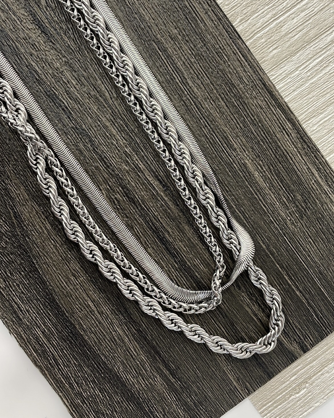 COLARES HOMEM quatro colares prateados com texturas diferentes sobre madeira escura