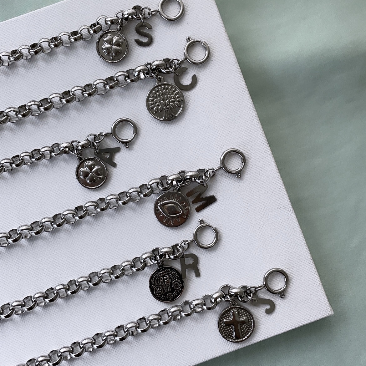 LUCK Cinco pulseiras metálicas prateadas com pingentes redondos e letras penduradas sobre superfície branca