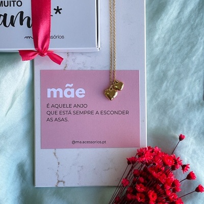 Colar dourado com coração sobre cartão rosa com texto e flores vermelhas em tecido azul.