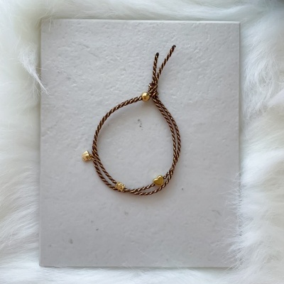 Pulseira de cordão castanho com detalhes dourados em forma de coração, concha e quadrado sobre base branca.
