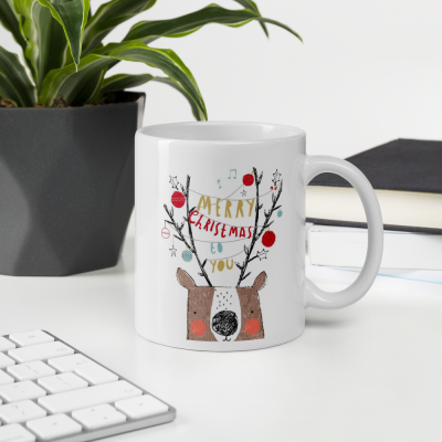 Caneca branca com desenho de alce e texto de Natal numa mesa com teclado, planta e livros