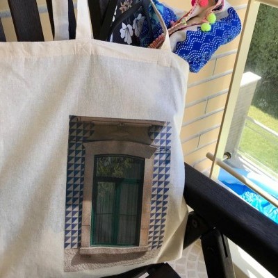 Saco de pano bege com imagem de janela verde e padrão de azulejos azuis pendurado numa cadeira preta numa varanda