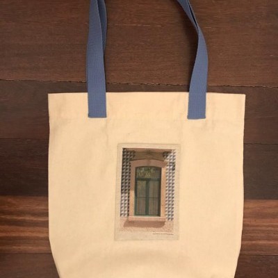 Saco de tecido bege com alças azuis e imagem de janela com azulejos.