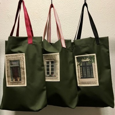 Três sacos de tecido verde-oliva com diferentes alças e imagens de portas ou janelas pendurados numa parede branca
