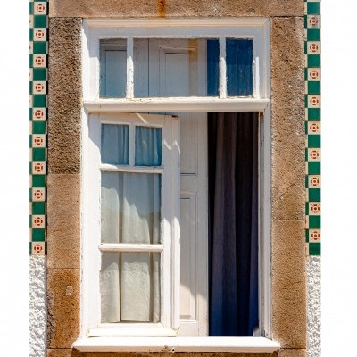 Janela branca de madeira com cortinas e moldura de pedra com azulejos decorativos verdes.