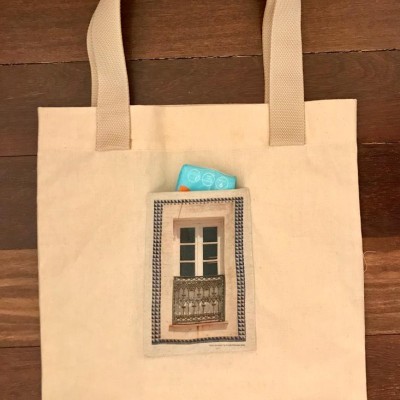 Saco de tecido bege com bolso e alças largas, com imagem de janela e objeto azul no bolso.