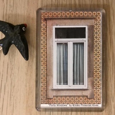 Íman com imagem de janela branca e cortinas claras, padrão decorativo castanho, ao lado de ornamento em forma de pássaro preto