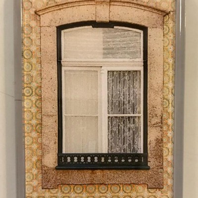 Janela emoldurada por pedra com azulejos amarelos de padrão circular e cortinas de renda brancas.