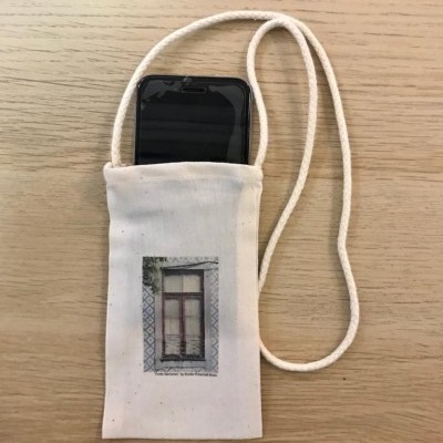 Bolsa de tecido branco com alça fina e imagem de janela antiga, telemóvel dentro, sobre superfície de madeira