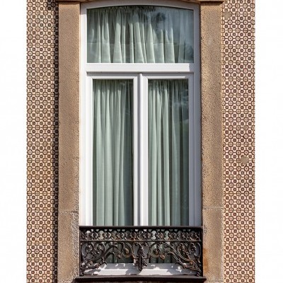 Janela branca com cortinas cinza, moldura de pedra bege, azulejos castanhos e varandim de ferro forjado preto
