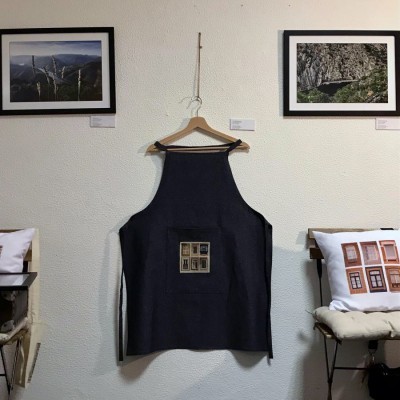 Avental preto com bolso estampado pendurado numa parede branca entre almofadas e fotografias emolduradas.