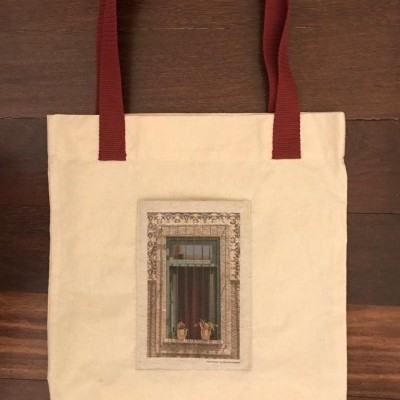 Saco tote creme com pegas vermelhas e imagem de janela com plantas.