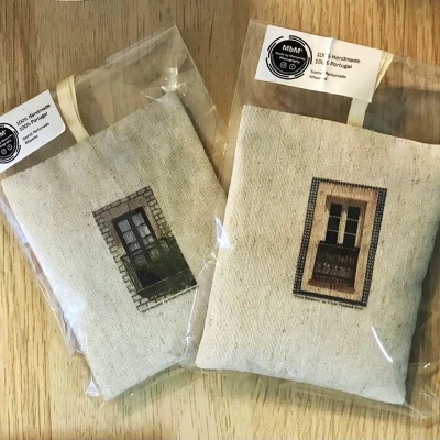 Dois sacos pequenos de tecido bege com imagens de janelas em mosaico, embalados em plástico com etiquetas brancas em fundo de madeira clara.
