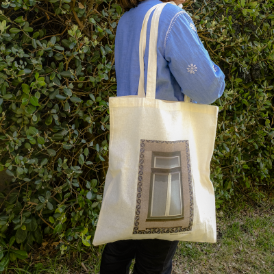 Mala tote de tecido bege com imagem impressa de porta, usada por pessoa de camisa azul em ambiente exterior