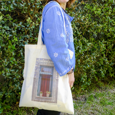 Sacola de algodão cru com estampa de janela, usada por pessoa com camisa azul bordada, em ambiente com vegetação.