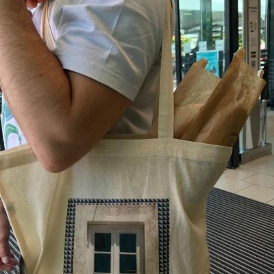 Saco de pano bege com impressão de janela azul, carregado por uma pessoa numa área interna.