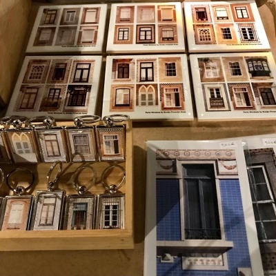 Porta-chaves e postais com imagens de janelas tradicionais em várias cores, mostrando castanho, branco, cinzento e azul.