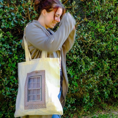 Mala tote de tecido bege com impressão de janela, alças largas, usada por pessoa com casaco castanho claro, em frente a sebe verde.