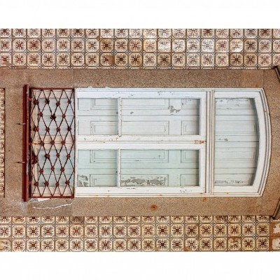 Janela branca de madeira com moldura de pedra e gradeado vermelho, em parede de azulejos decorativos.