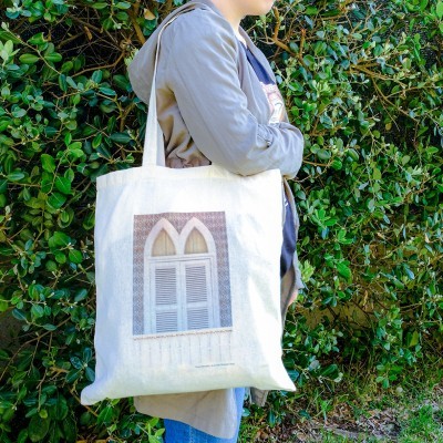 Saco de tecido branco com impressão de janela arqueada, usado por pessoa junto a folhagem verde.
