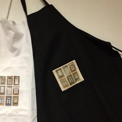 Dois aventais, branco e preto, com estampas de janelas coloridas, pendurados na parede branca