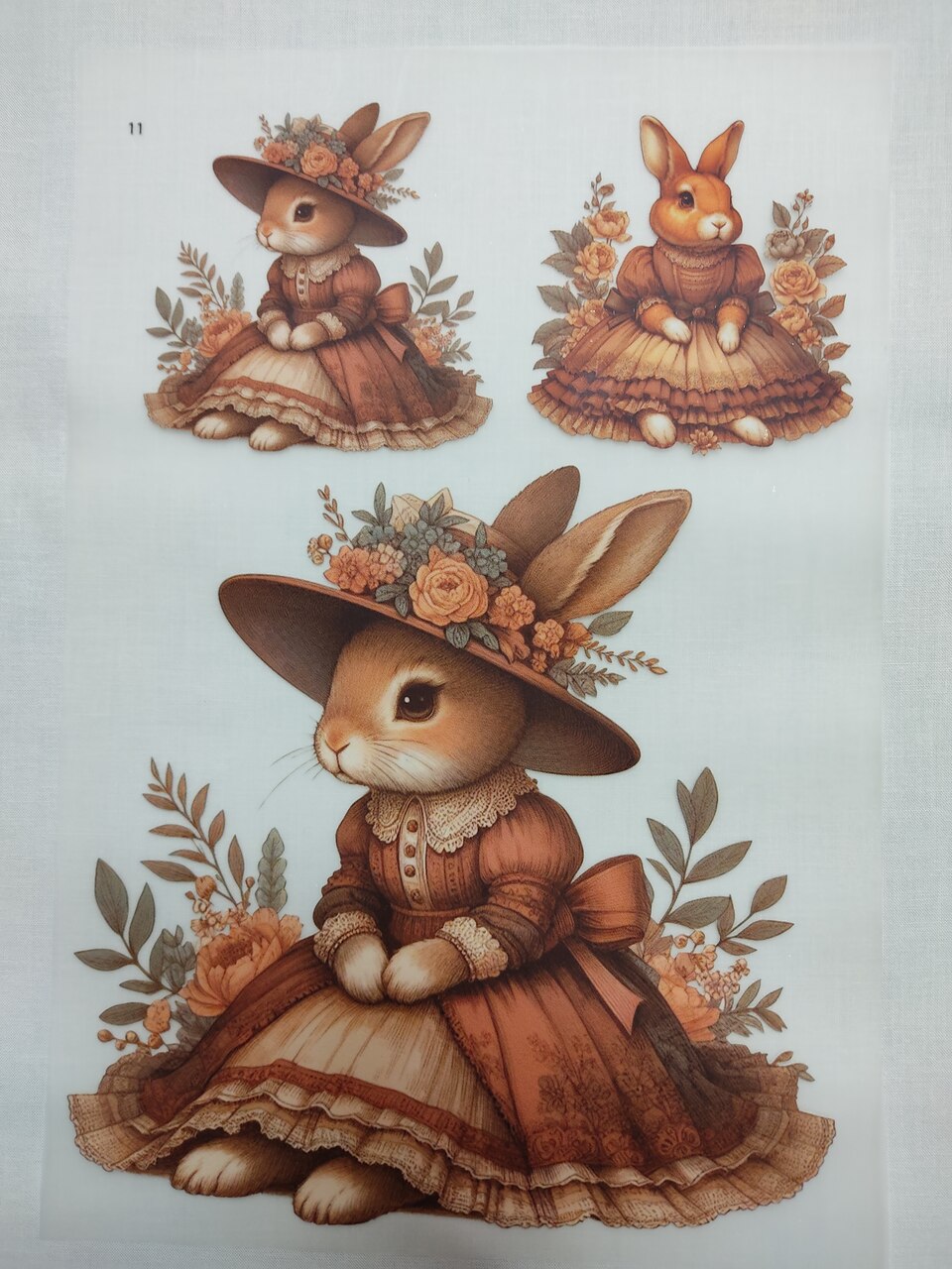 Transfere "Coelha com chapéu e flores" DTFR 11 Tecido estampado com coelhos vestidos em roupa antiga cor-de-rosa e castanho com chapéus floridos.